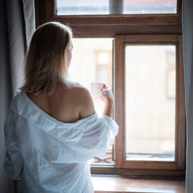 Zarif bornozlu kadın otel odasında kahve içiyor ve pencerenin yanında duruyor. Pencere yansıması