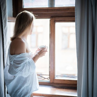 Zarif bornozlu kadın otel odasında kahve içiyor ve pencerenin yanında duruyor. Pencere yansıması