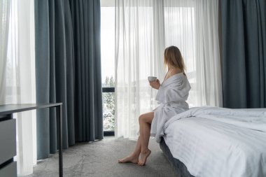 Woman in white bathrobe lying on sofa and relaxing with cup of tea at home near window