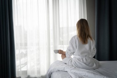 Woman in white bathrobe lying on sofa and relaxing with cup of tea at home near window