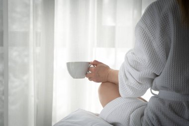 Woman in white bathrobe lying on sofa and relaxing with cup of tea at home near window