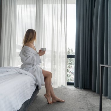 Woman in white bathrobe lying on sofa and relaxing with cup of tea at home near window