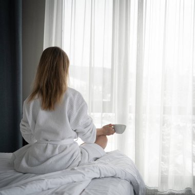 Woman in white bathrobe lying on sofa and relaxing with cup of tea at home near window
