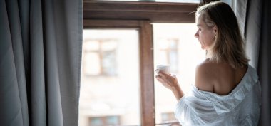 Sabahleyin otelin penceresinde beyaz havluyla duran genç bir kadın.