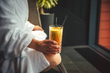 Woman in white bathrobe lying on sofa and relaxing with orange cocktail at home near window