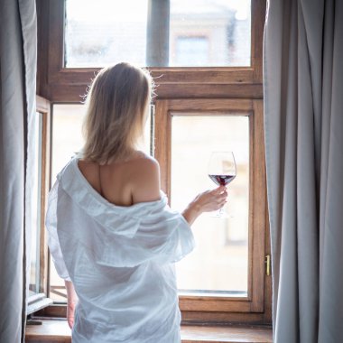 Oteldeki yatak odasında elinde bir kadeh kırmızı şarap tutan kadının arka görüntüsü.