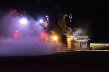 Une moissonneuse-batteuse s'affaire dans un champ de bl la nuit tombe. claire par ses puissantes lampes, elle avance lentement en digrant les pis dors qui se dressent devant elle. 