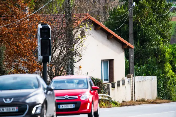 La photo montre une rota avec des voitures on mouvement devant une maison. Il a un radar de vitesse sur le cote de la route, des arbres feiilles caduques et des arbustes verts convert sonble autour. 