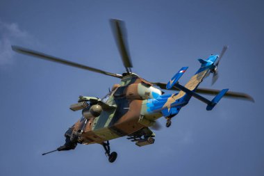 Bir Fransız askeri Tigre helikopteri havada, kamuflaj ve mavi işaretlerle süslenmiş, açık gökyüzünde dönen pervanelerle. Helikopter gelişmiş silah ve gözetleme sistemleriyle donatılmıştır..