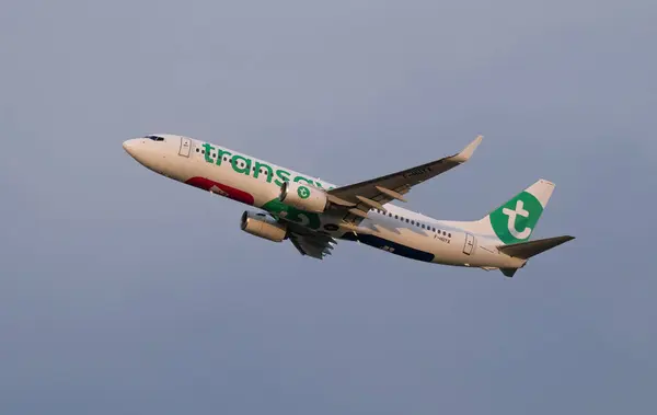 Yeşil ve beyaz üniformalı bir Transavia yolcu uçağı açık bir gökyüzüne yükselir. Uçağın motorları, uçağın gövdesinde belirgin bir şekilde markalaşmasını gösteren yolculuğuna çıkarken kükrüyor..