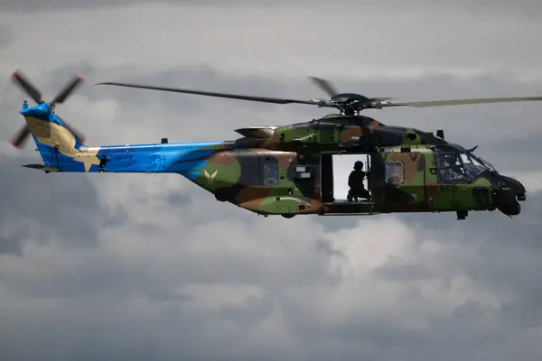 Kamuflaj boyası ve büyük pervane bıçaklarıyla uçan bir NH90 Caiman TTH helikopteri. Askerler açık kapıdan görülebiliyor, askeri ulaşım ve operasyonlardaki rolünü vurguluyorlar.