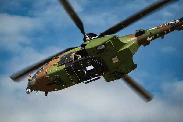 Fransız Ordusu AS-332 Cougar helikopteri, yeşil ve kahverengi kamuflaj modelini gösteriyor. Helikopter parçalı bulutlu bir gökyüzüne doğru süzülüyor ve askeri ulaşım ve operasyonlardaki rolünü vurguluyor..