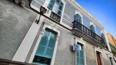 Demir balkon ve mavi panjurlu tarihi bina açık gökyüzüne vurgulanarak ayrıntılı tasarımını gözler önüne seriyor.