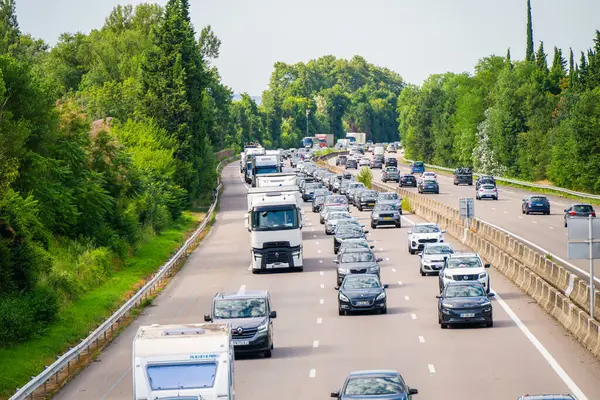 Otoyol sahnesi yoğun trafiği gösteriyor. Yeşillikler arasında çeşitli araçlar iki yönde ilerliyor..