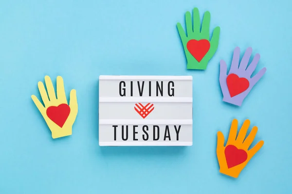 Giving Tuesday, global day of charitable giving after Black Friday shopping day. Charity, give help, donations support concept. Colorful hands, white lightbox, red heart on blue background. Top view, copy space, flat lay.