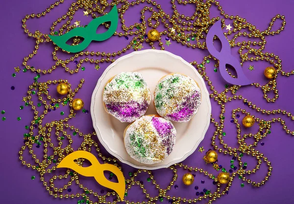 Mardi Gras King Cake sufganiyot donuts, masquerade festival carnival masks, gold beads and golden, green, purple confetti on purple background. Holiday party invitation, greeting card concept.