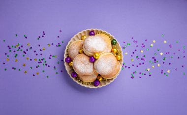 Mardi Gras King Cake doughnuts or donuts, masquerade festival carnival mask, gold beads and golden, green confetti on purple background. Holiday party invitation, greeting card concept.