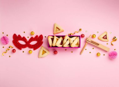 Purim celebration jewish carnival holiday concept. Tasty hamantaschen cookies, Triangular pastry, red carnival mask, noisemaker, sweet candies and party decor on pink background. Top view, flat lay, copy space.
