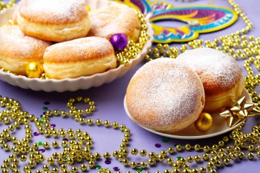 Mardi Gras King Cake doughnuts or donuts, masquerade festival carnival mask, gold beads and golden, green confetti on purple background. Holiday party invitation, greeting card concept.