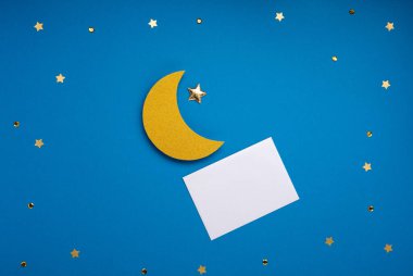 Ramadan Kareem holiday concept. Golden crescent moon, stars and empty white card on blue background. Traditional Muslim Iftar. Top view, copy space.