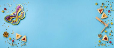 Purim celebration jewish carnival holiday concept. Tasty hamantaschen cookies, Carnival mask, noisemaker, sweet candies and party decor on blue background. Top view, flat lay, copy space.