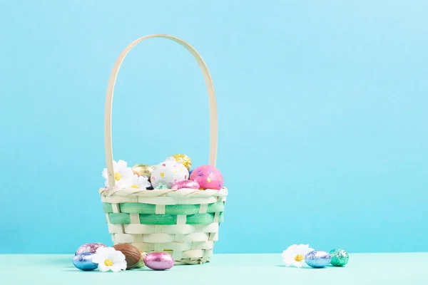 Easter Basket, Sweet Colorful Chocolate Easter Eggs, daisy flowers on pastel blue mint background. Happy Easter greeting card, Egg hunt concept, copy space.