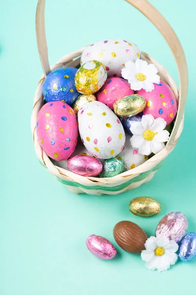 Easter Basket, Sweet Colorful Chocolate Easter Eggs, daisy flowers on pastel blue mint background. Happy Easter greeting card, Egg hunt concept, copy space.