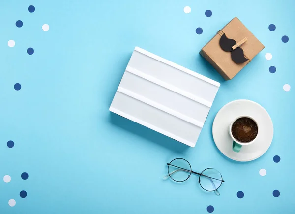 Fathers day holiday card concept with craft gift box, black moustache, cup of coffee, empty white lightbox and glasses on blue background, top view, copy space.