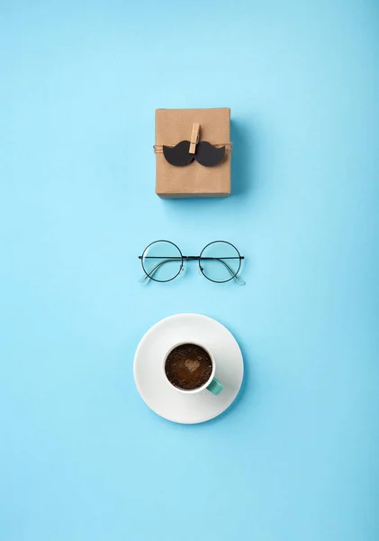 Fathers day holiday card concept with craft gift box, black moustache, cup of coffee and glasses on blue background, top view, copy space.
