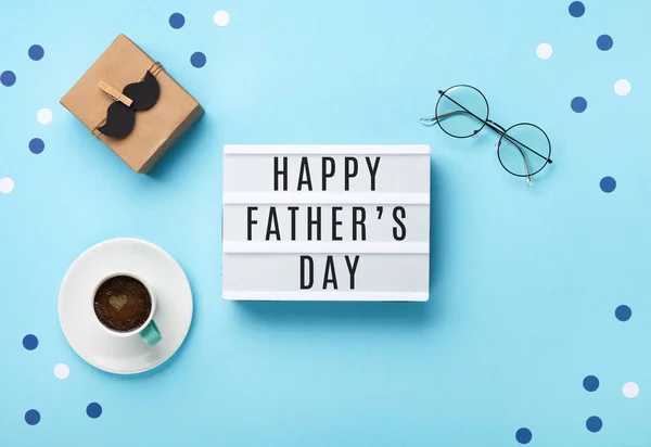 Fathers day holiday card concept with craft gift box, black moustache, cup of coffee, empty white lightbox and glasses on blue background, top view, copy space.