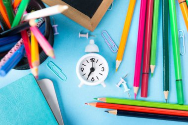 Okula, sınava, mavi arka planda eğitim konseptine. Okul dağınık kırtasiye malzemeleri, not defterleri, kalemler, ataçlar ve mavi masadaki beyaz çalar saat. Üst görünüm, alanı kopyala. 