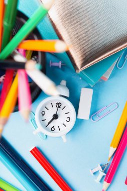Okula, sınava, mavi arka planda eğitim konseptine. Okul dağınık kırtasiye malzemeleri, not defterleri, kalemler, ataçlar ve mavi masadaki beyaz çalar saat. Üst görünüm, alanı kopyala. 
