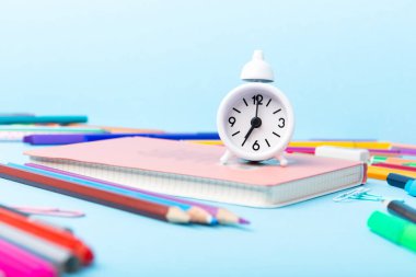 Okula, sınava, mavi arka planda eğitim konseptine. Okul dağınık kırtasiye malzemeleri, not defterleri, kalemler, ataçlar ve mavi masada beyaz alarm saati, kopyalama alanı. 