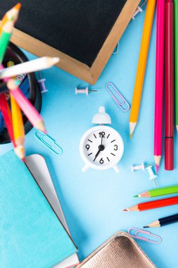 Okula, sınava, mavi arka planda eğitim konseptine. Okul dağınık kırtasiye malzemeleri, not defterleri, kalemler, ataçlar ve mavi masadaki beyaz çalar saat. Üst görünüm, alanı kopyala. 