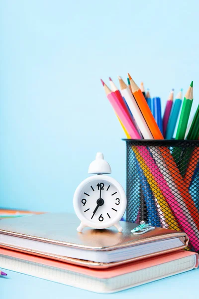 Back to school, Exam, Education concept on blue background. School scattered stationery, notepads, pens, pencils, paper clips and white alarm clock on blue table, copy space.