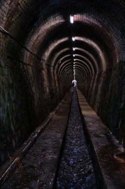 Uzun bir tünel, yapay ışıklarla aydınlatılmış, duvarlarla çevrili, su kanalı.