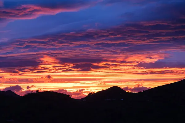 stock image A vibrant sunset casts a warm glow over the sky, with fluffy clouds drifting above towering mountains in the background. The suns rays create a stunning display of colors on the horizon.