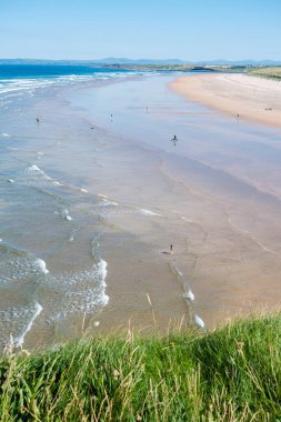 Sörfçüler İrlanda 'nın Donegal kentindeki Playa en Finner' da dalgaların tadını çıkarıyorlar. Geniş kumlu sahil, parlak mavi bir gökyüzünün altında uzanır. Rahatlamaya davet eden sakin bir atmosfer yakalar..