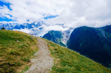Sarmal bir yol yemyeşil çimlerin arasından geçer, arka planda görkemli Alpler yükselir. Gökyüzü parçalı bulutlu ve bu güzel manzaranın sakin atmosferine ekleniyor..