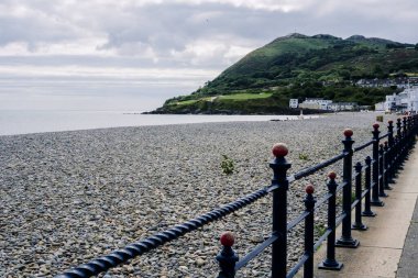 Bray, İrlanda 'da sakin bir çakıl taşı plajı kıyı şeridi boyunca uzanır, yeşil tepeler ve bulutlu gökyüzü altında dağınık beyaz kulübelerle çerçevelenir, barışçıl bir geri çekilme için mükemmel.