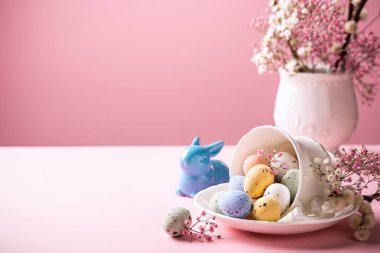 Easter composition with spring flowers and colorful quail eggs in porcelain white coffee cup over pink background. Springtime and Easter holiday concept with copy space