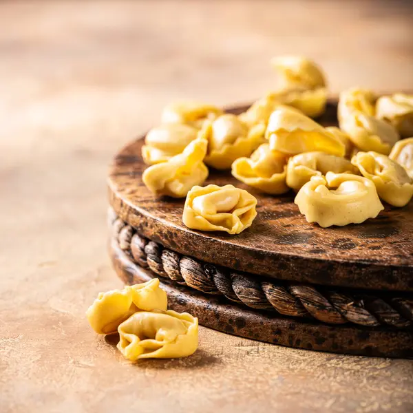 Köy tahtasının üzerinde pişmemiş tortellini makarna, fotokopi alanı olan sıcak desenli bir arka plan üzerine kurulmuş. Sağlıklı İtalyan yemeği konsepti