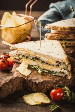 Taze malzemelerle dolu istiflenmiş sandviç, ahşap bir tahtada çıtır cips ve vişneli domateslerle servis ediliyor, enfes bir mutfak deneyimi vurgulanıyor.