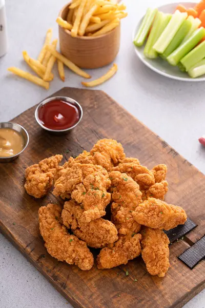 Chicken nuggets or chicken strips on a board with dipping sauces, fries and celery sticks