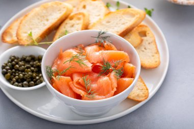 Dilimlenmiş füme somon tabağı dereotuyla süslenmiş, kızarmış baget dilimleri ve kapari ile eşleştirilmiş.