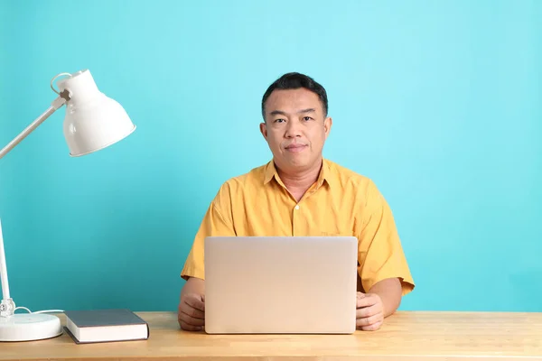 四十多岁的亚洲男子 身穿黄色衬衫 坐在蓝色背景的办公桌前 — 图库照片