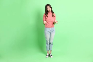 Yeşil arka planda duran, sıradan giyinmiş, kot pantolonlu genç Asyalı kadın..