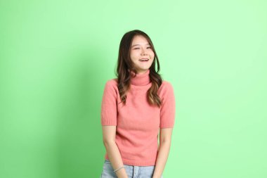 Yeşil arka planda duran, sıradan giyinmiş, kot pantolonlu genç Asyalı kadın..