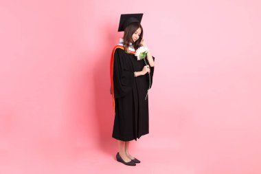 Mezuniyet elbisesi giymiş, pembe arka planda duran Asyalı kadın..