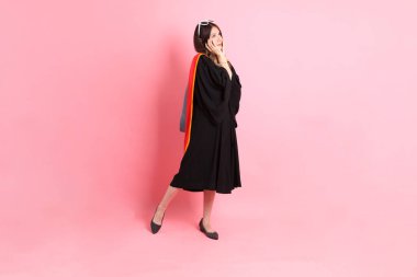 Mezuniyet elbisesi giymiş, pembe arka planda duran Asyalı kadın..